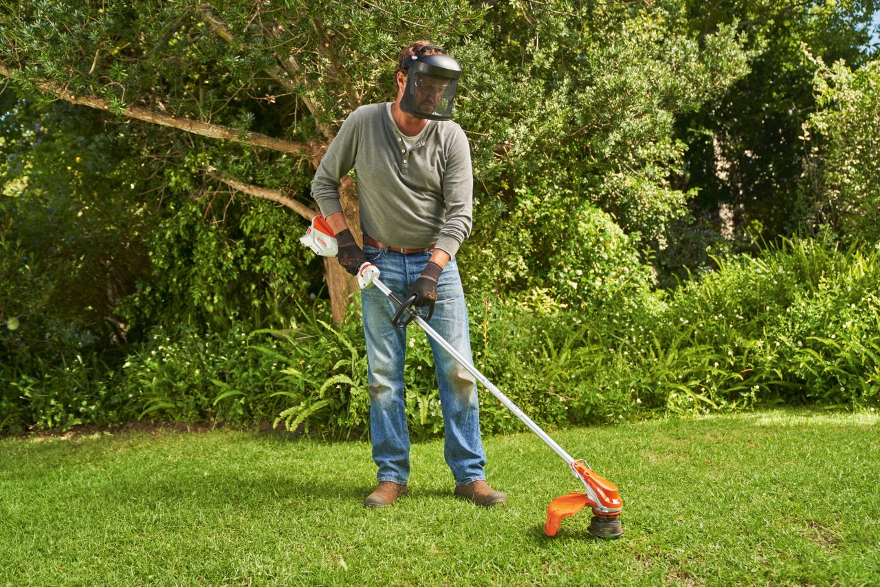 顔面保護具を着用して、STIHL FSA 60バッテリー刈払機で庭の芝刈りをしようとしている男性